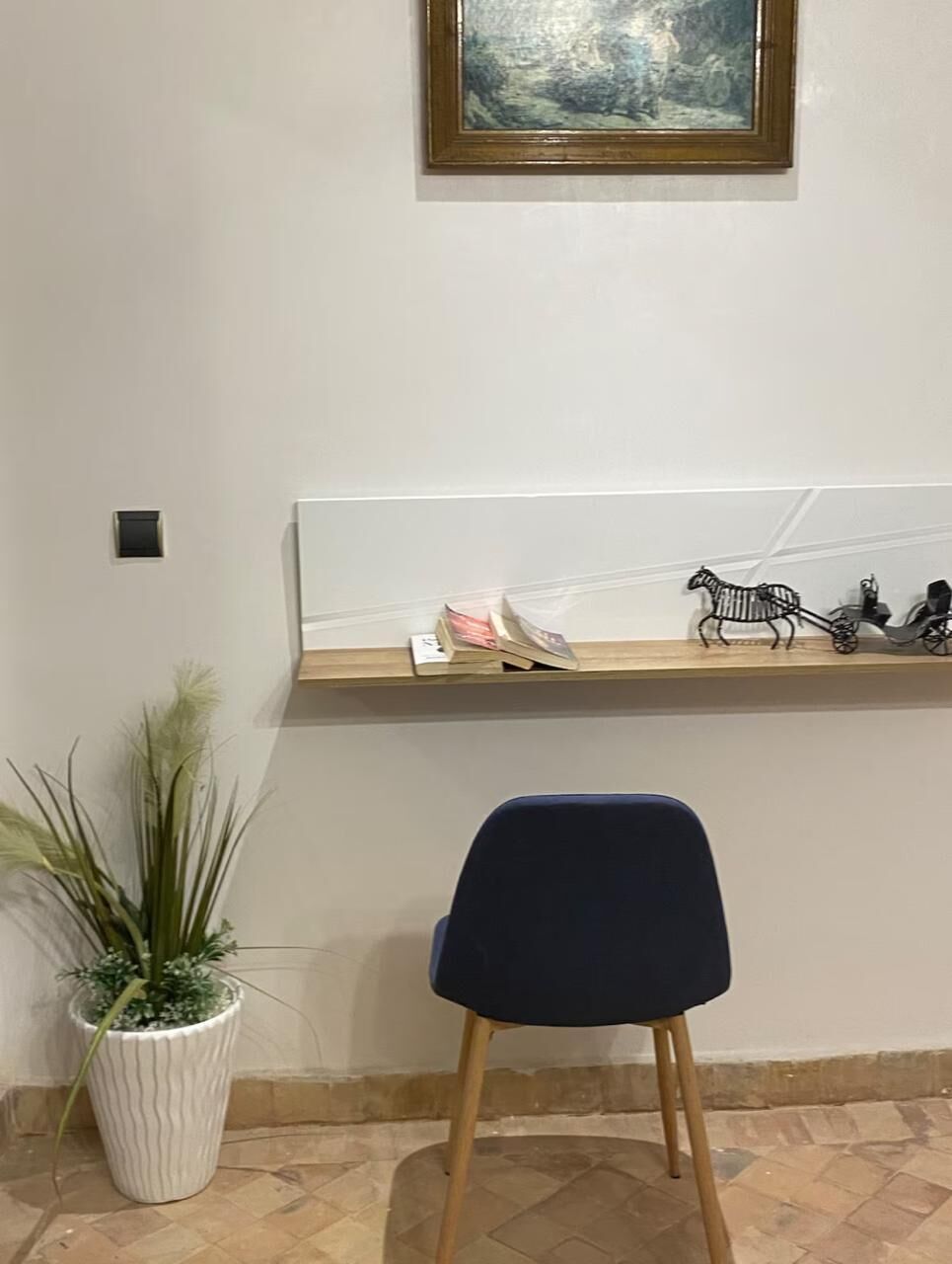 A room with a white wall, a framed painting, a shelf with decorative items, a potted plant, and a dark blue chair.
