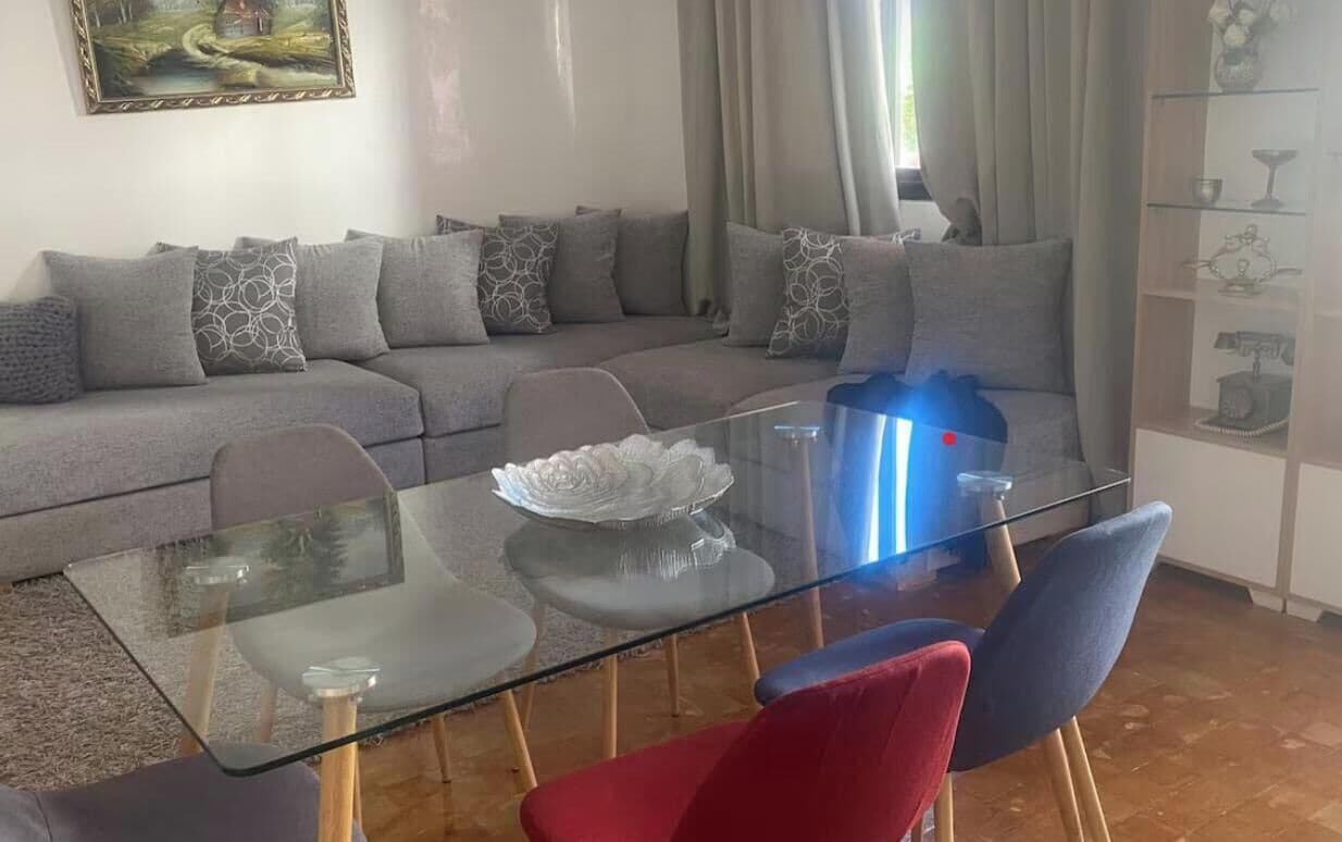 A living room with a glass coffee table, a sofa with patterned cushions, and a sideboard with decorative items.