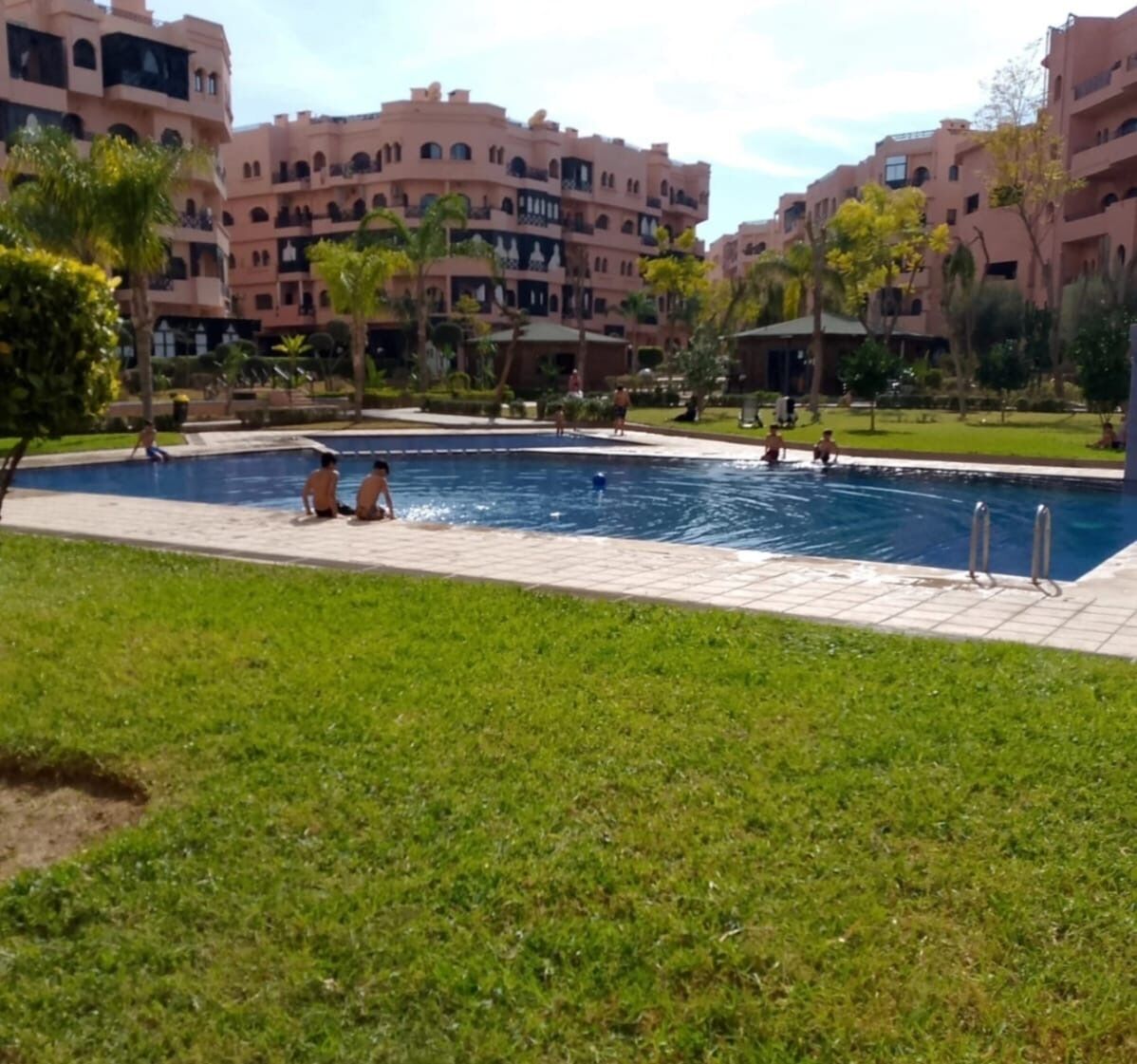 Une zone avec piscine où se trouvent plusieurs personnes, entourée d’une pelouse verdoyante et de bâtiments en arrière-plan.