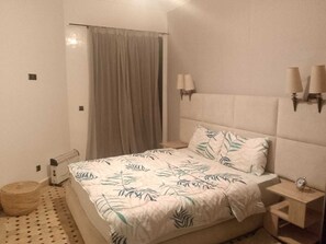 Ein Schlafzimmer mit einem Bett, einem Nachttisch, einer Lampe, einer Wandleuchte, einem Vorhang und einem gemusterten Boden.