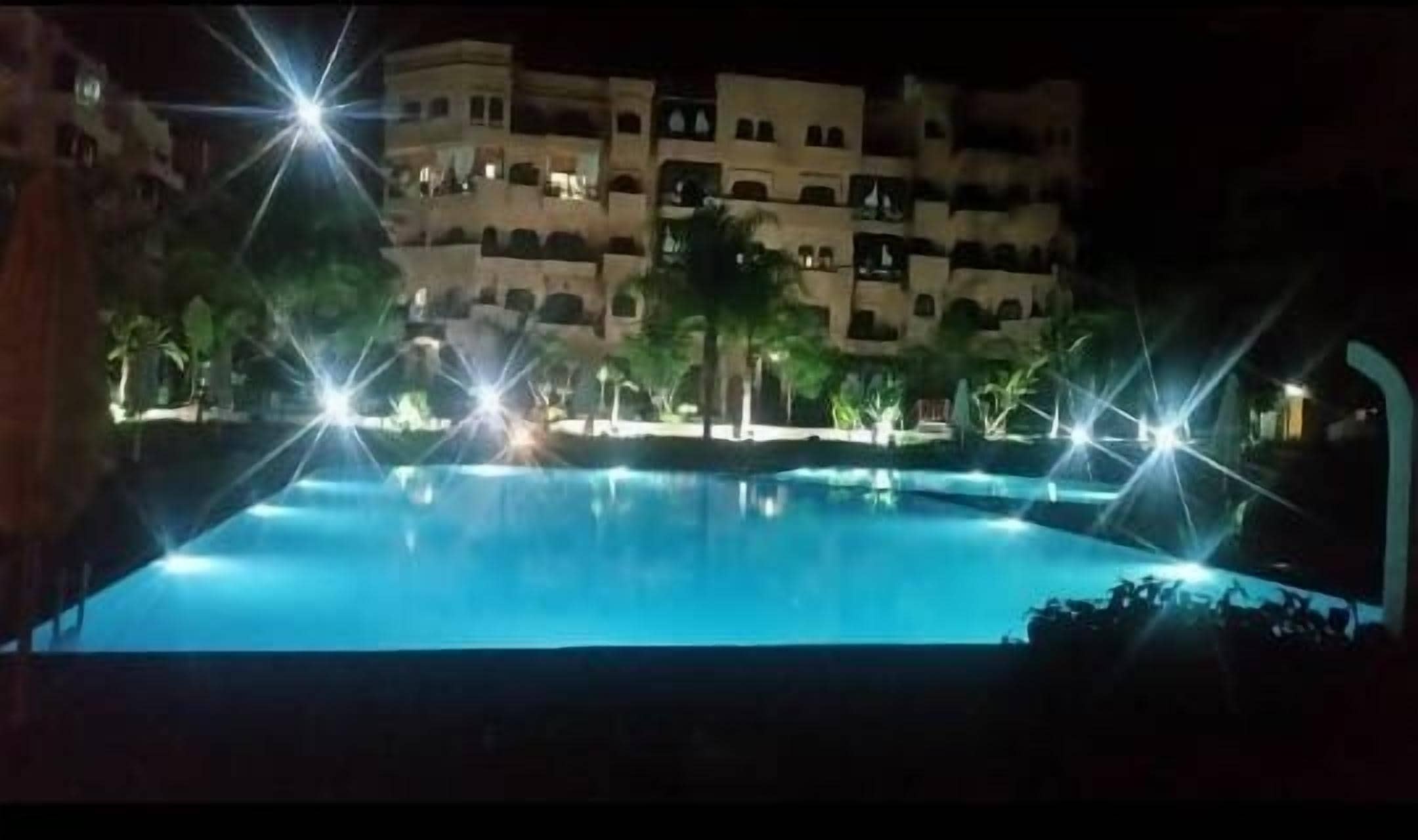 Ein Nachtblick auf einen Pool mit umliegenden Palmen und einem Gebäude mit mehreren Balkonen.