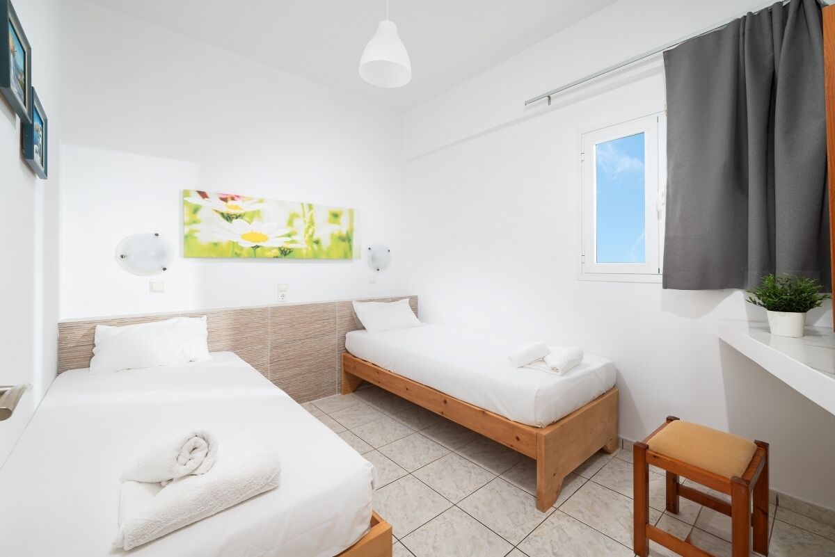 A modern hotel room with two single beds, a wooden bench, a small table, a potted plant, and a window with curtains.