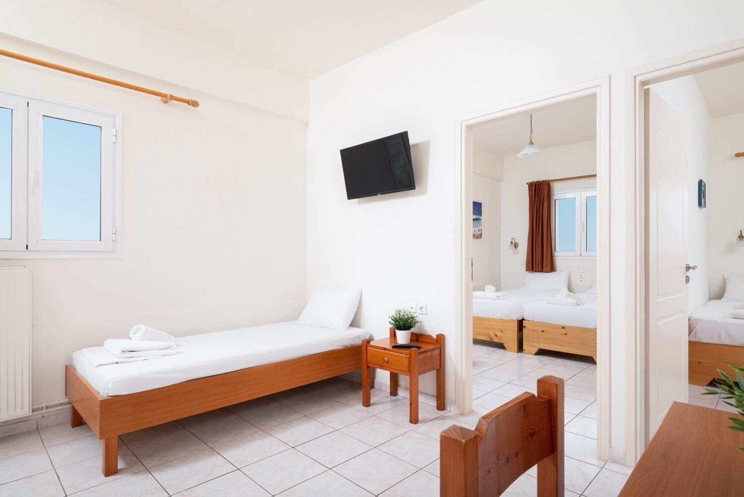 A single-bed room with a wooden bed, a small bedside table with a plant, a TV mounted on the wall, and a view into an adjacent room with a bed and a window.