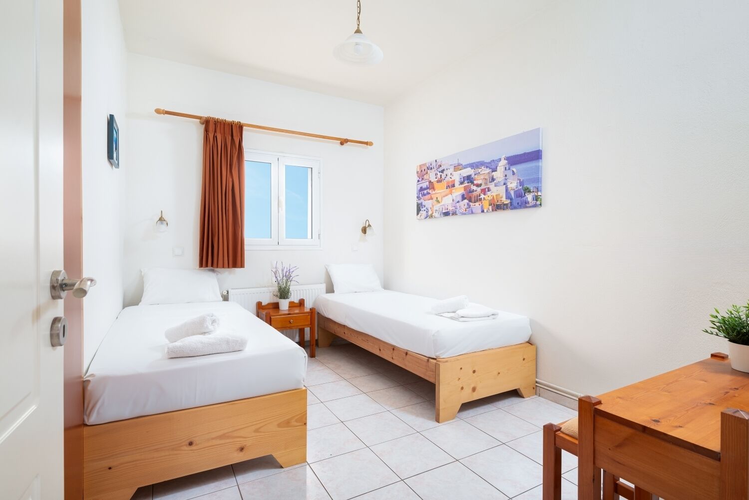 A room with two beds, a small table, a chair, a potted plant, a window with curtains, and a painting on the wall.