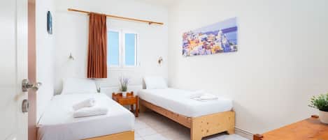 A room with two beds, a small table, a chair, a potted plant, a window with curtains, and a painting on the wall.