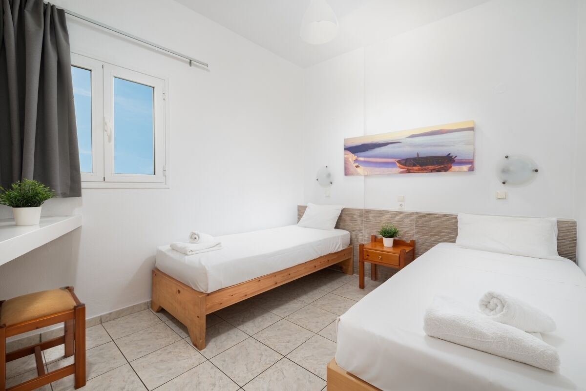 A hotel room with two single beds, a small table, a chair, a window with curtains, a wall-mounted painting, and a potted plant.
