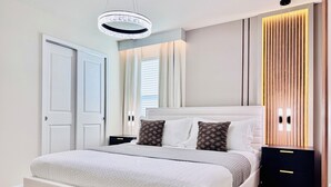 A modern bedroom with a large bed, a bedside table, a nightstand, a wall-mounted light fixture, and a wooden panel accent. - Spacious 7BR Clermont Retreat w/ 4 King Beds, Modern Comfort & Great Location (Clermont)