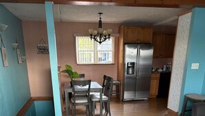 Een woonkamer met een grijze bank, een eethoek met een tafel en stoelen, een keuken met houten kastjes en een roestvrijstalen koelkast, en een kroonluchter.