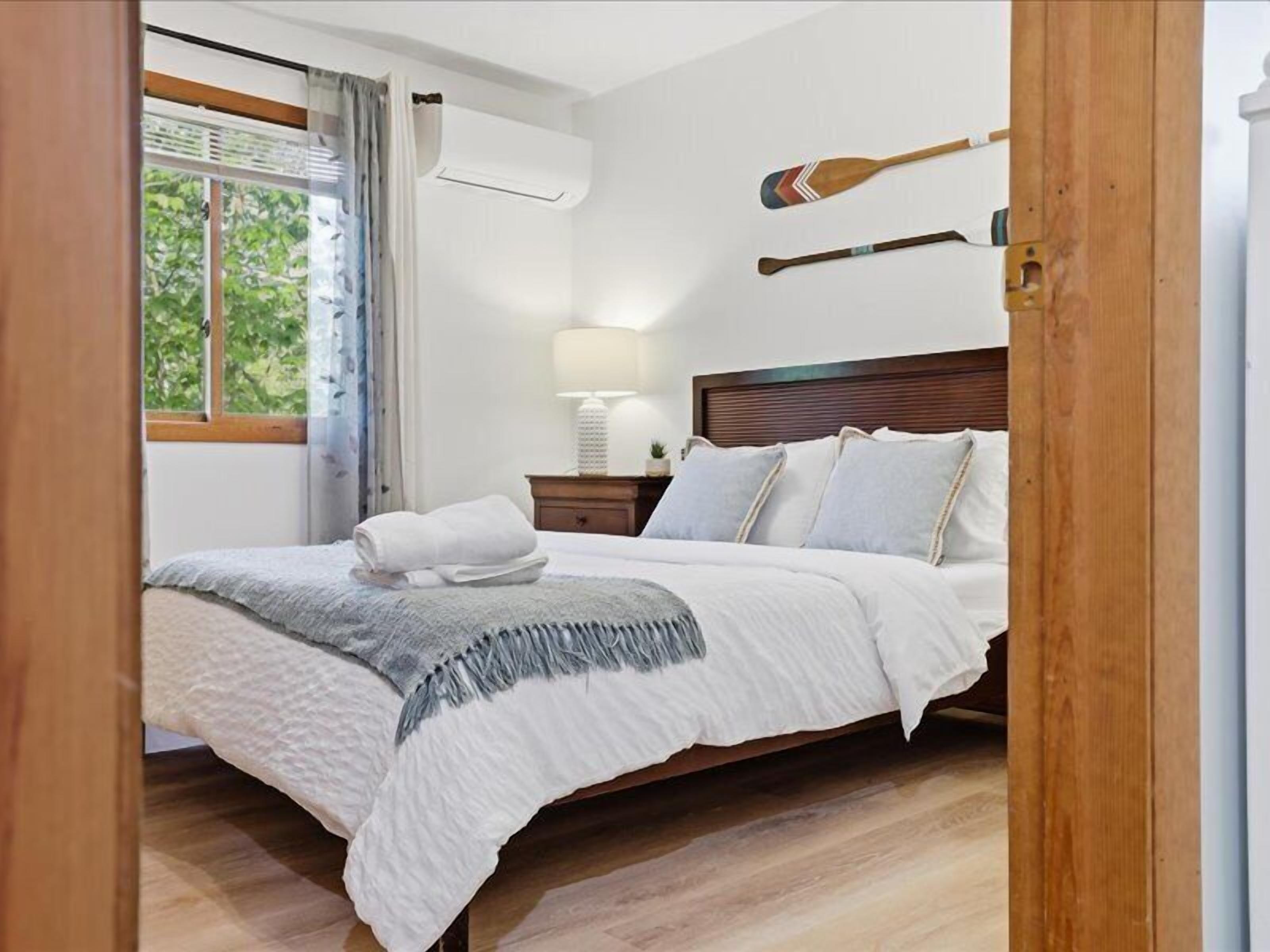 Ein ordentlich bezogenes Bett mit weißer Decke und Kissen, eine Nachttischlampe und zwei Holzschilder an der Wand.