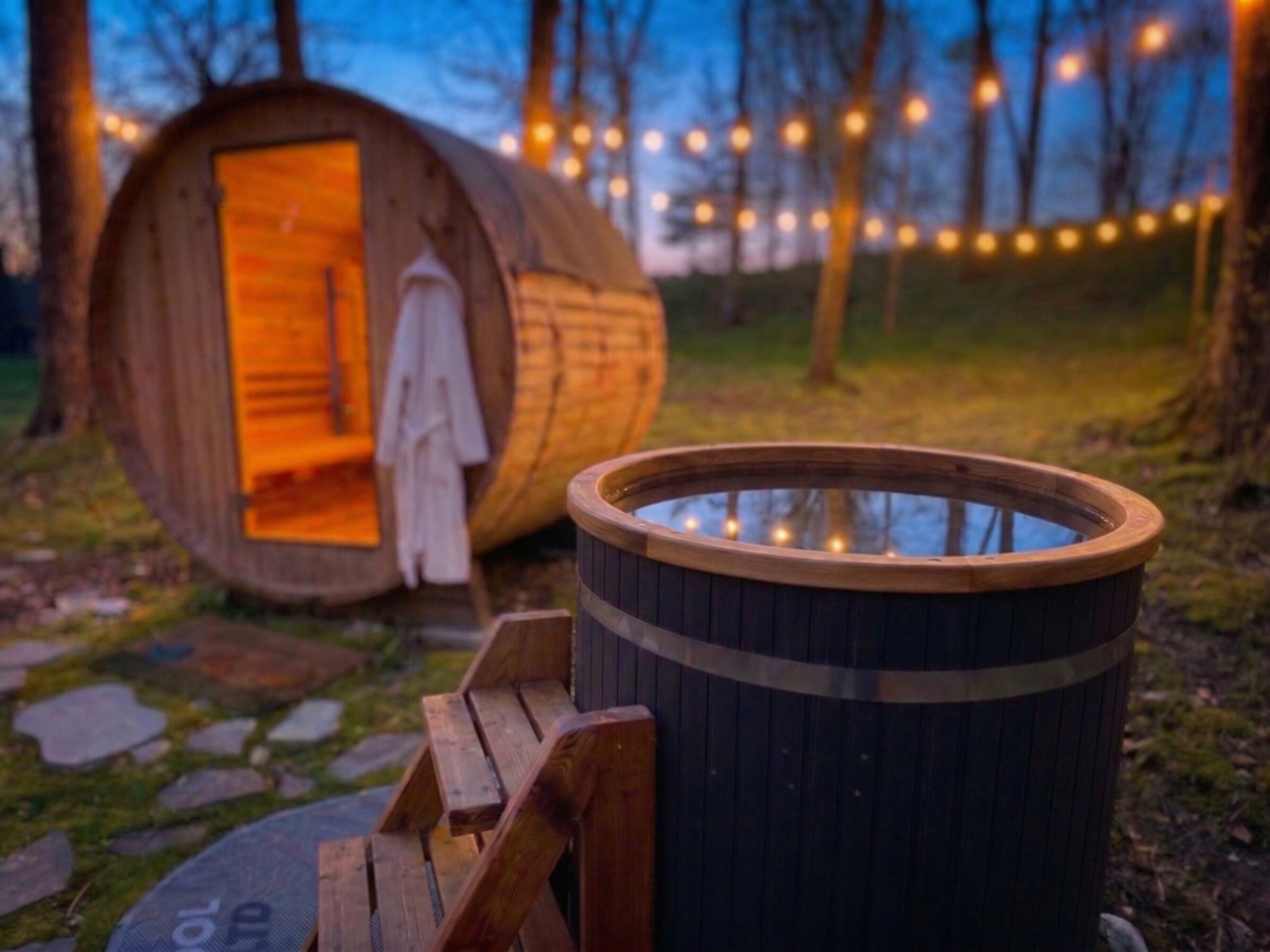 Eine Holzbottensauna mit Sitzbank davor, umgeben von einem Steinweg und einer Holzhütte mit offener Tür.