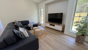 A modern living room with a sectional sofa, a flat-screen TV on a wooden stand, and a large window with a view of a wooden fence.