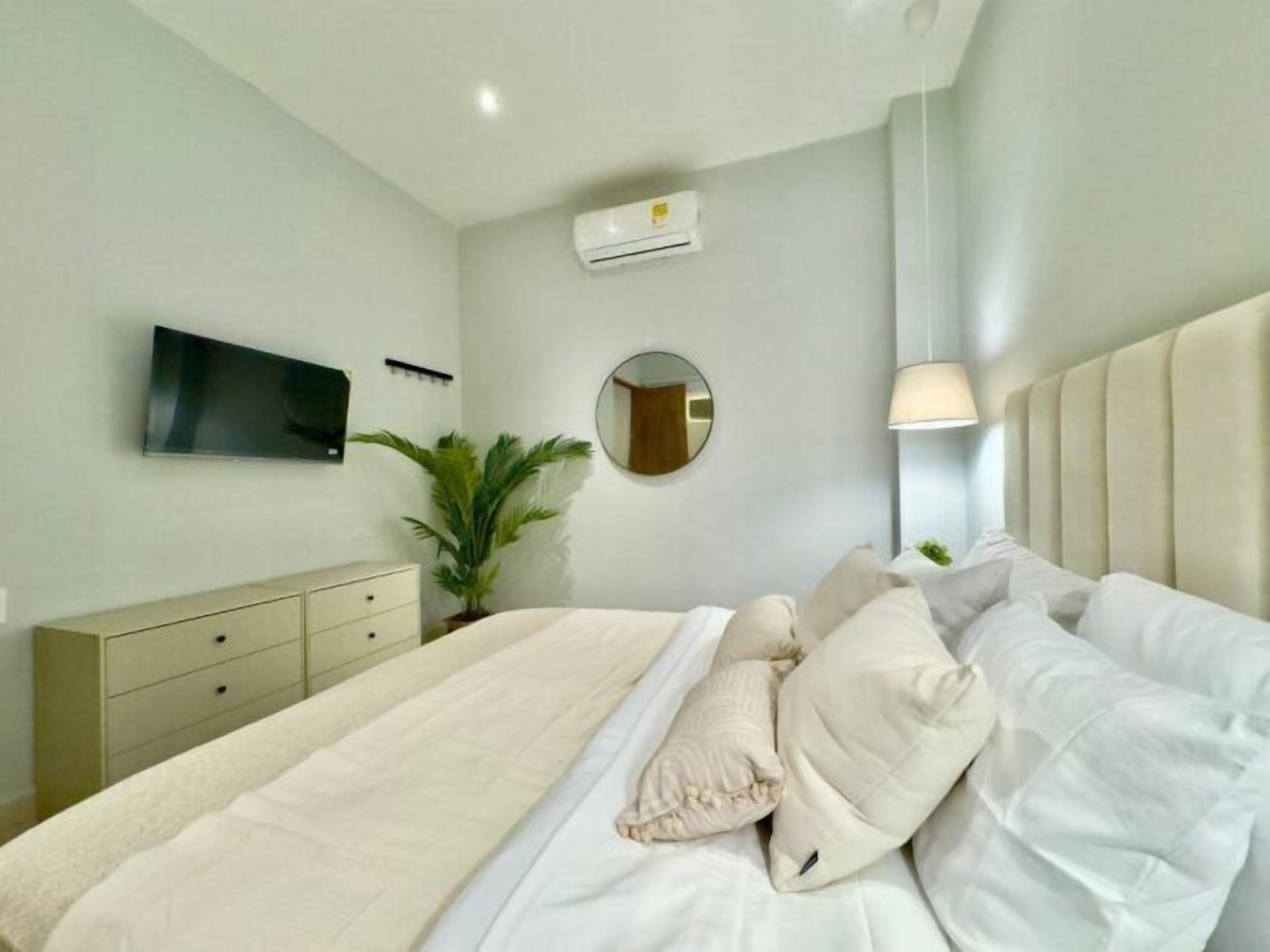 Une chambre à coucher moderne avec un lit, un téléviseur fixé au mur, un miroir rond et une plante en pot.