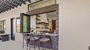 Un área de comedor al aire libre con pared de piedra y sillas de metal, una puerta de cristal que da a una cocina y una pérgola en lo alto.