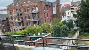 A balcony with a glass table and chairs, overlooking a brick building and greenery.