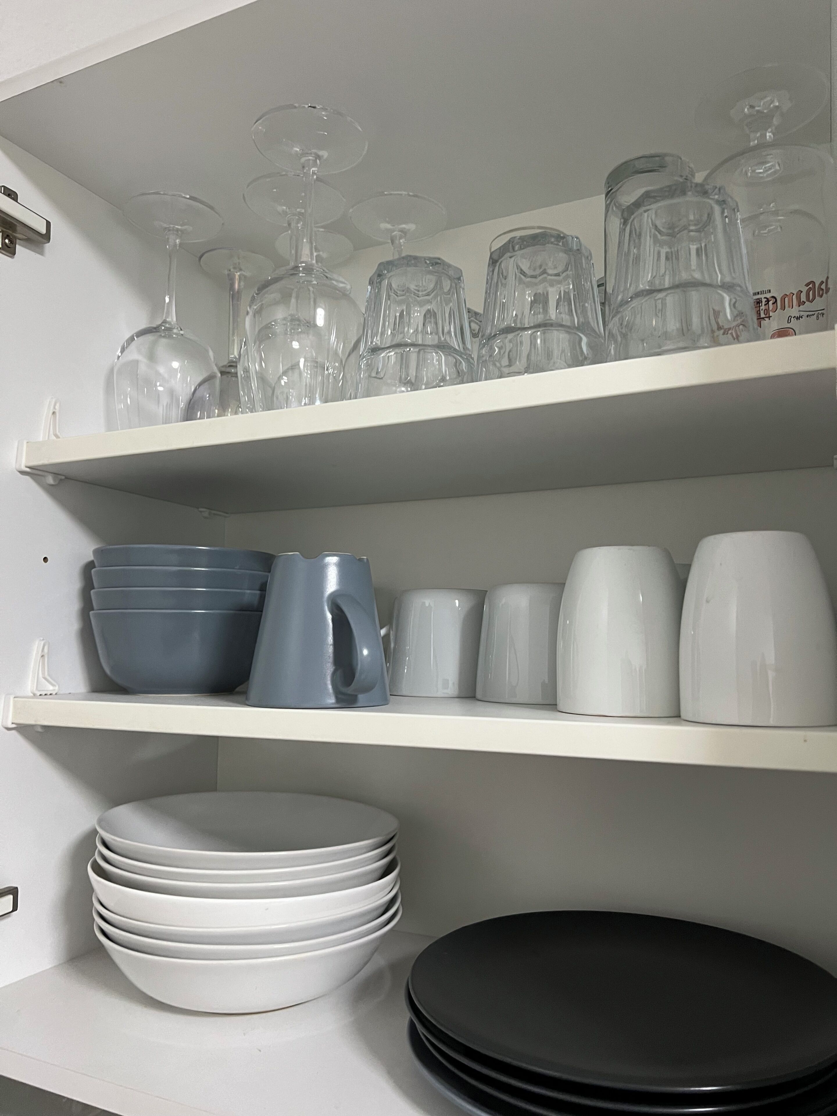 Una repisa de cocina con diversos vasos, incluyendo copas de vino y vasos sencillos, además de una pila de platos y cuencos blancos.