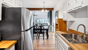 A modern kitchen with white cabinets, a stainless steel refrigerator, a wooden dining table with chairs, and a view of the outdoors through a large window.