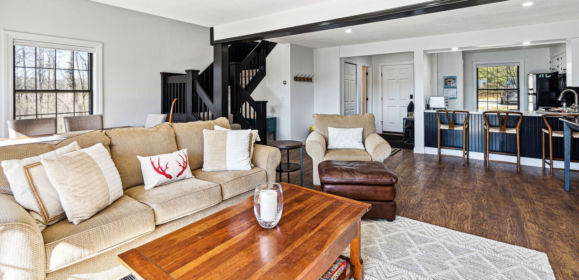 A modern living room with a wooden coffee table, a beige sofa with decorative pillows, a leather ottoman, and a staircase leading to another level.