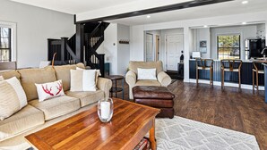 A modern living room with a wooden coffee table, a beige sofa with decorative pillows, a leather ottoman, and a staircase leading to another level. - Condo with Fireplace, Sugar Hill (Quechee)