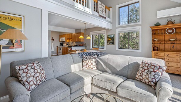 A spacious living room with a grey sectional sofa, a dining area, and a kitchen. - Ski-In/Ski-Out Condo with Big Back Deck, Kingswood (Quechee)