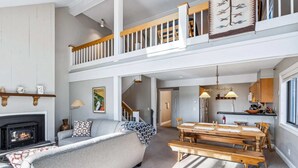 A spacious living room with a fireplace, a sofa, a dining table, and a staircase leading to an upper level. - Ski-In/Ski-Out Condo with Big Back Deck, Kingswood (Quechee)