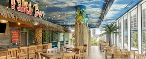 Un restaurant avec des meubles en bois, un toit de chaume et une fresque représentant un paysage tropical.