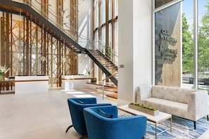Un hall d’hôtel moderne avec un comptoir d’accueil, des fauteuils bleus, un escalier et une grande fenêtre donnant sur l’extérieur.