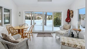 Ein heller, geräumiger Raum mit einer großen Schiebeglastür, die zu einer verschneiten Außenanlage führt. Im Inneren steht ein Holzespeisetisch mit Stühlen, ein Rattansofa und ein kleiner Beistelltisch mit einer Lampe.