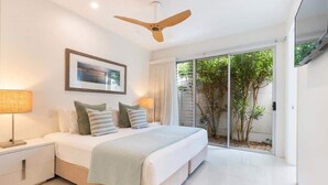 A modern bedroom with a large bed, a ceiling fan, a framed picture on the wall, and a sliding glass door leading to a garden.