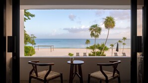 A balcony with two wooden chairs and a small table, overlooking a beach with palm trees and a bench.