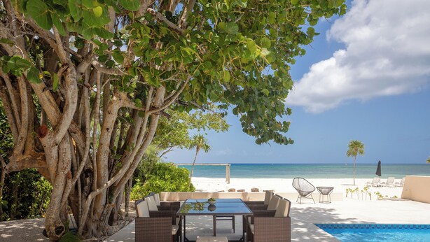 A poolside area with a dining table and chairs, a view of the beach, and a large tree.
