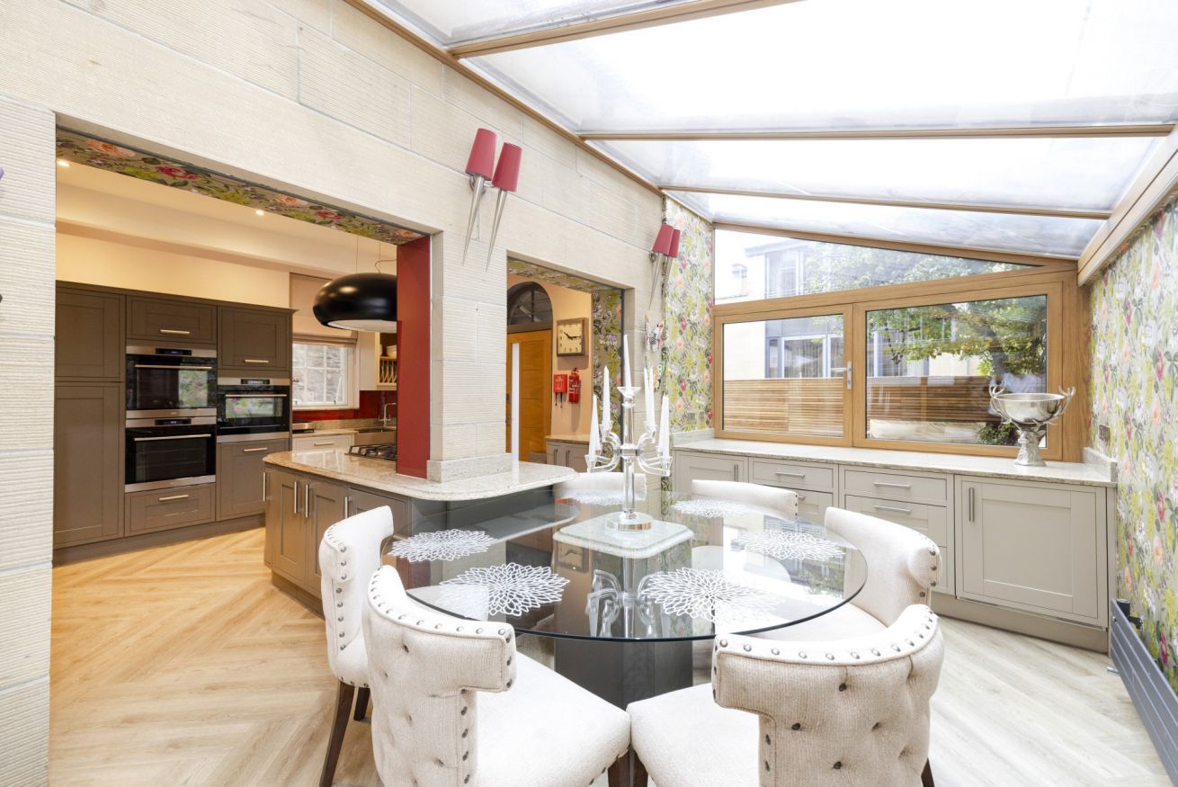 Une cuisine moderne dotée d’un îlot central, d’un coin repas avec une table en verre et offrant une vue sur l’extérieur grâce à de grandes fenêtres.