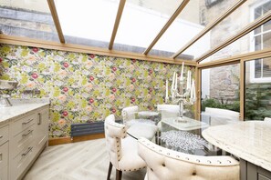 A conservatory with a floral wallpaper, a round glass table, and a white upholstered chair.