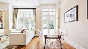 A modern living room with a sofa, a dining table with chairs, large windows, and a framed picture on the wall.