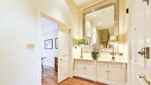 A hallway with a wooden floor, white walls, a large mirror, and a white console table with a lamp and vase.