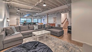A modern living room with a sectional sofa, a coffee table, and a large rug. There is a staircase leading to another level, and a dining area with a table and chairs. - Chain of Lakes Escape with Hot Tub & River Access! (Bellaire)