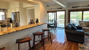 A modern kitchen with a granite countertop, wooden stools, and a large mirror. There is a dining area with a table and chairs, and a living room with a sofa and a large window. - Chain of Lakes Escape with Hot Tub & River Access! (Bellaire)