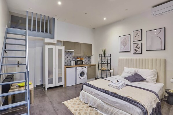 A bedroom with a bed, a small desk, a washing machine, a staircase, and a kitchen area.