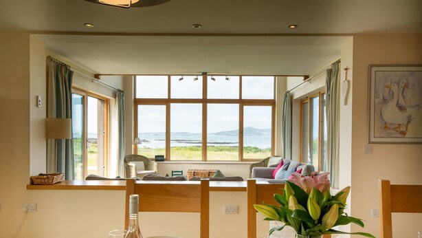 Een eetruimte met een tafel voor twee personen, een vaas met bloemen en uitzicht op de zee door grote ramen.