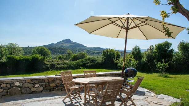 An outdoor patio with a stone table and chairs, a large umbrella, and a grill.