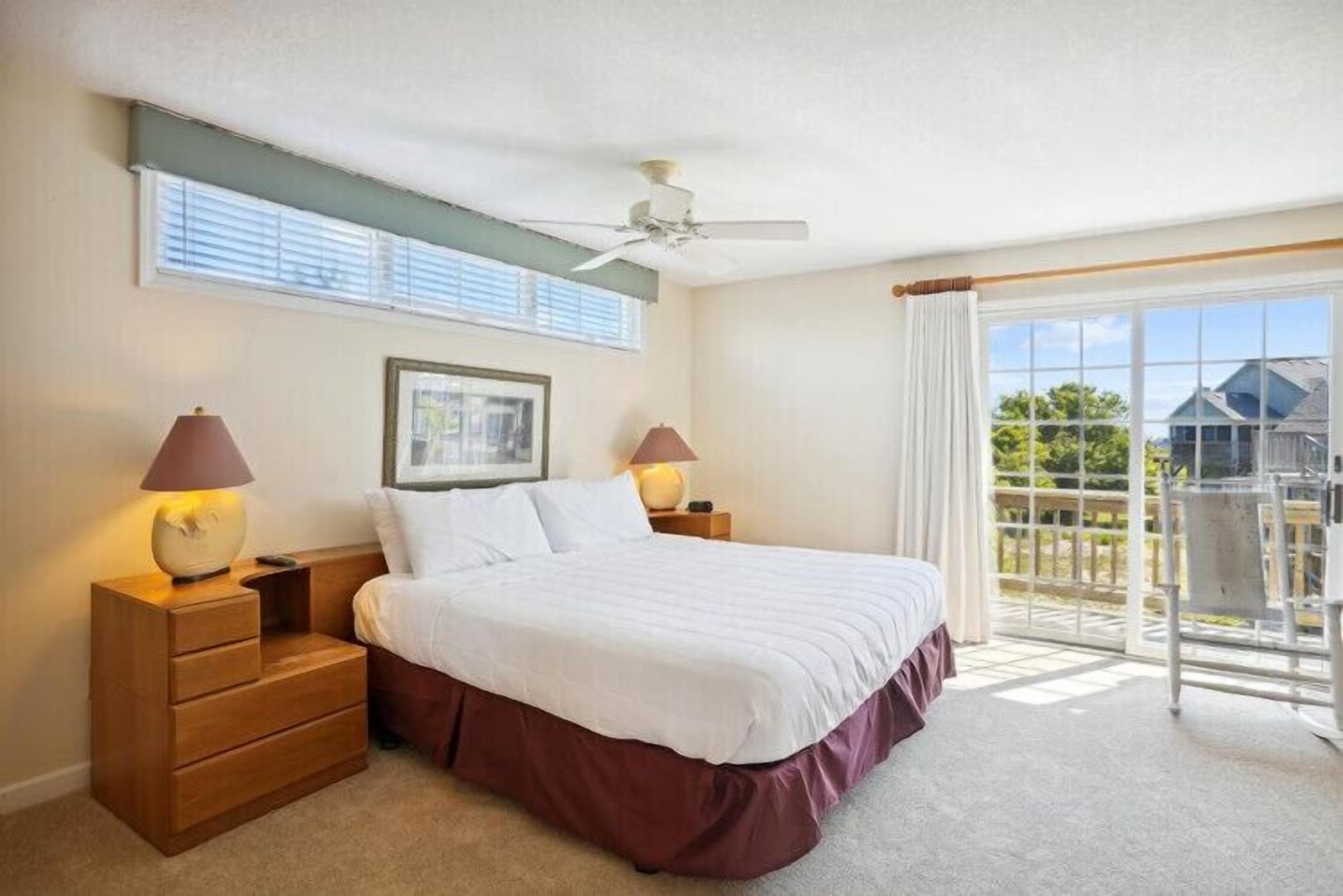 A bedroom with a bed, nightstands, a ceiling fan, and a view of the outside.