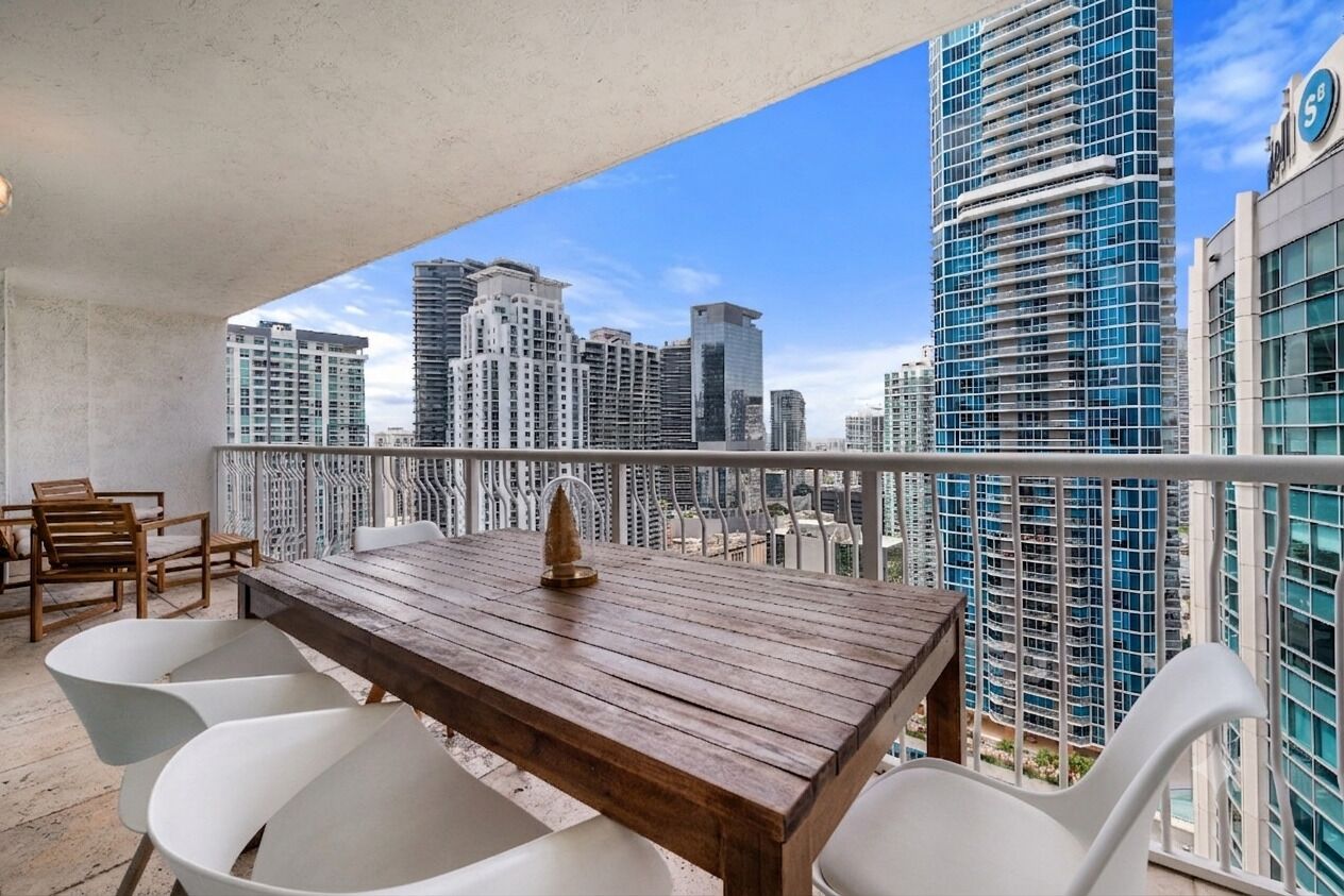 A balcony with a wooden table and white chairs, overlooking a cityscape with high-rise buildings.