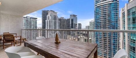 A balcony with a wooden table and white chairs, overlooking a cityscape with high-rise buildings.