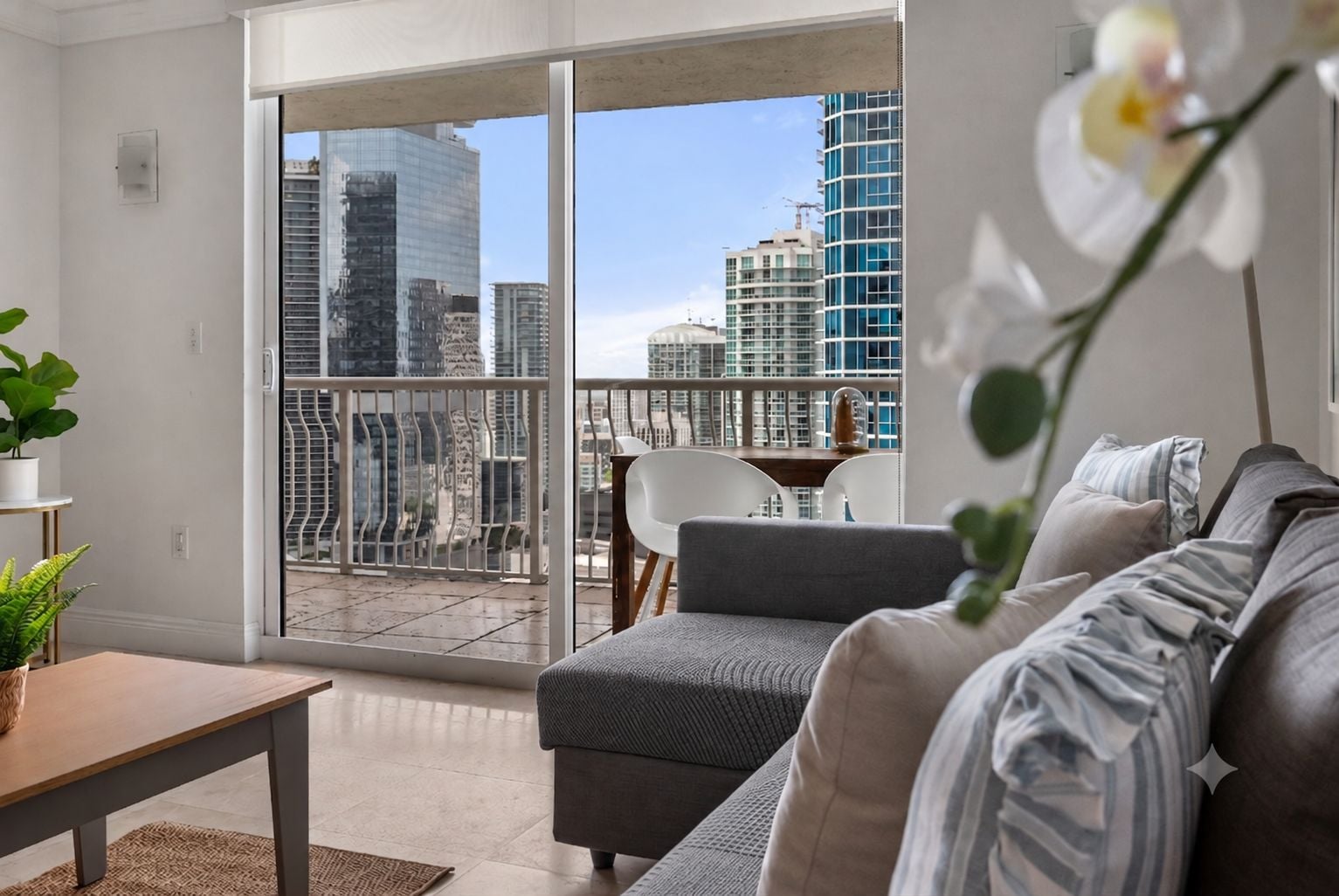 Un soggiorno moderno con un divano grigio, un tavolino in legno e una pianta in vaso. Un balcone con una porta vetrata offre vista sulla città.