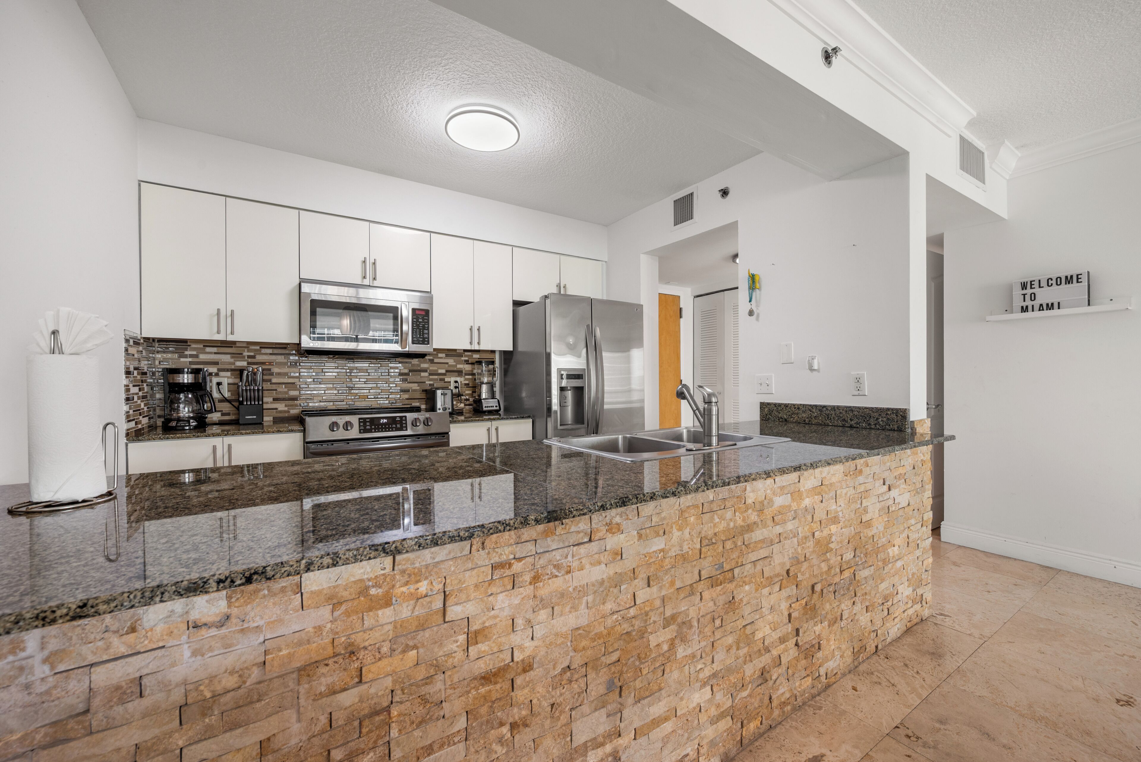 A modern kitchen with white cabinets, a granite countertop, a brick accent wall, stainless steel appliances, and a 'WELCOME TO MIAMI' sign.
