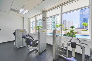 Une salle de sport équipée de machines d’exercice, dotée de grandes fenêtres et offrant une vue sur des immeubles et une piscine.