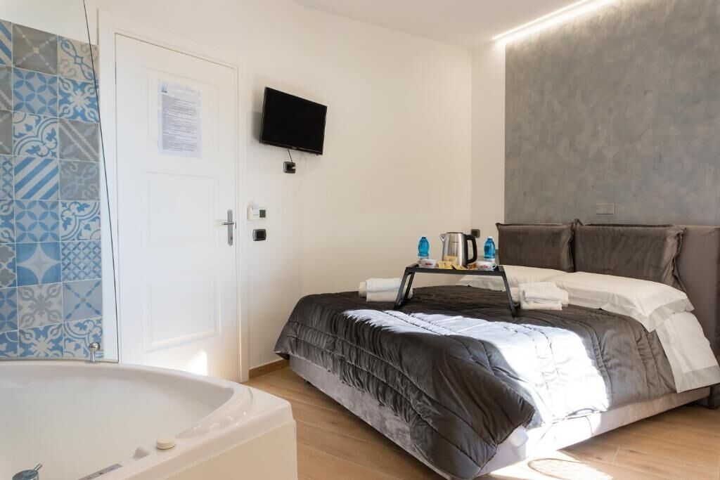 A bedroom with a bed, a TV mounted on the wall, a bedside table with toiletries, and a bathroom with a bathtub.