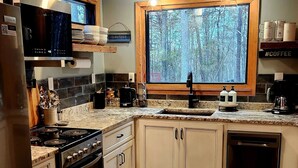 Une cuisine moderne dotée d’une cuisinière, d’un four et d’un lave-vaisselle intégrés. Elle comprend une grande fenêtre offrant une vue sur les arbres, un plan de travail en bois et un pare-feu composé d’un mélange de carreaux.