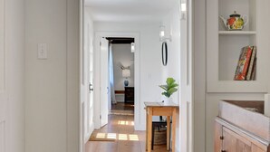 A hallway with wooden flooring, a small table with a plant, a wall-mounted lamp, and a shelf with books and a teapot.