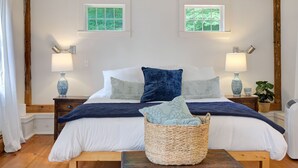 A bedroom with a wooden ceiling, two windows, a bed with white and blue bedding, two bedside tables with lamps, and a wicker basket on the floor.