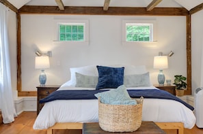 A bedroom with a wooden ceiling, two windows, a bed with white and blue bedding, two bedside tables with lamps, and a wicker basket on the floor.